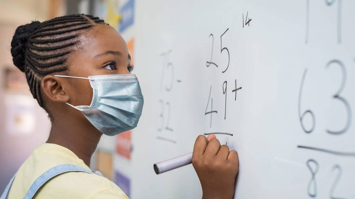 Young Black girl wearing face mask and writing solution of sums on white board at school