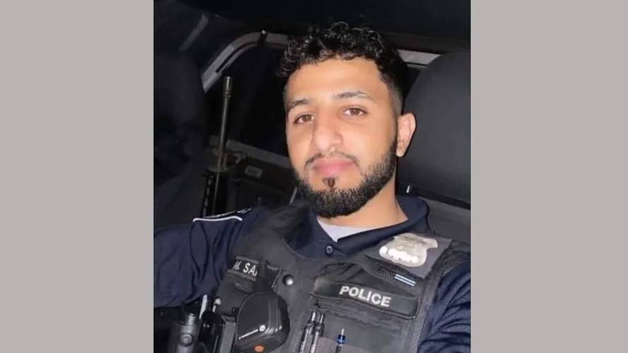 Young police officer with beard, wearing tactical vest & 'POLICE' patch, in car.